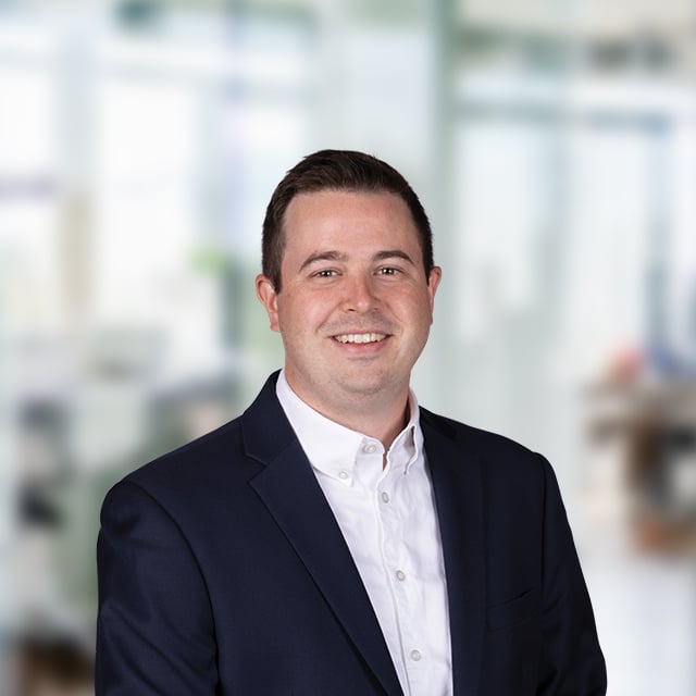 A man in a suit smiles at the camera, with a blurred office background.