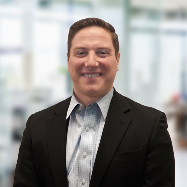 Man in a blazer and open collared shirt smiling with a blurred office background.