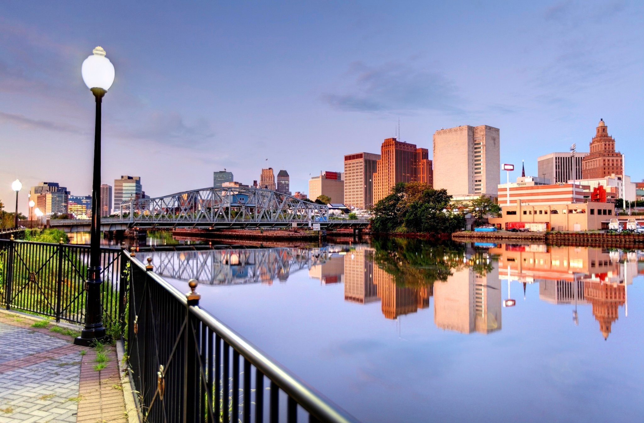 City buildings reflected in water, bridge, lamp post in view.