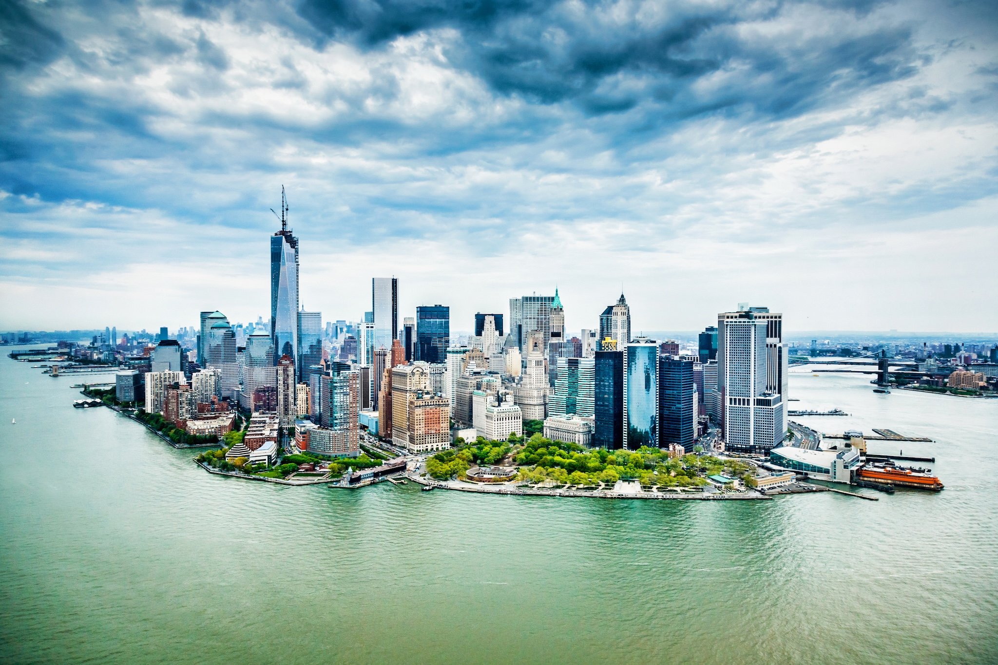 New York City skyline, cloudy sky, water in foreground. Skyscrapers and buildings.