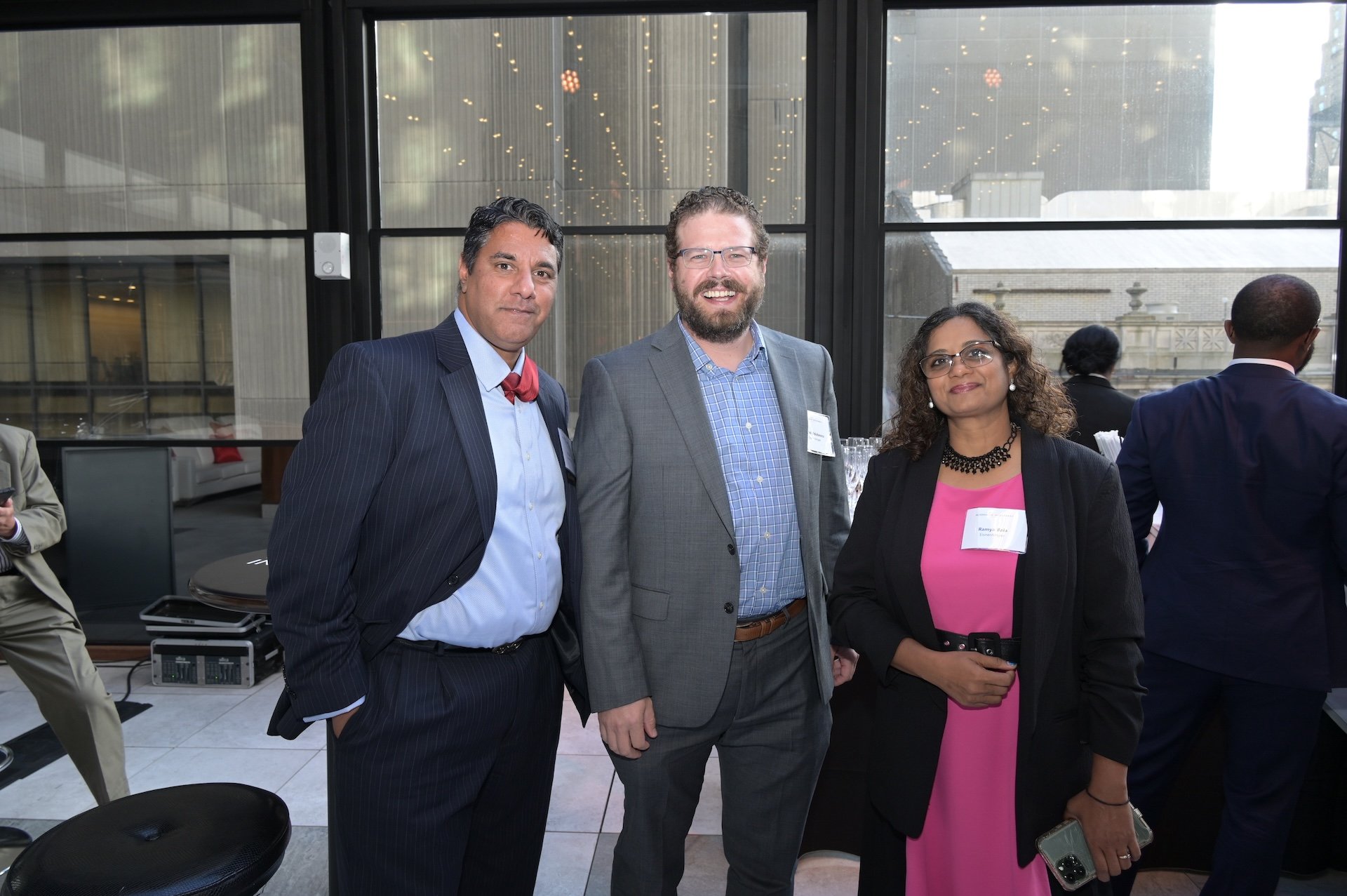 Three people smiling at event. Woman in pink dress and glasses.