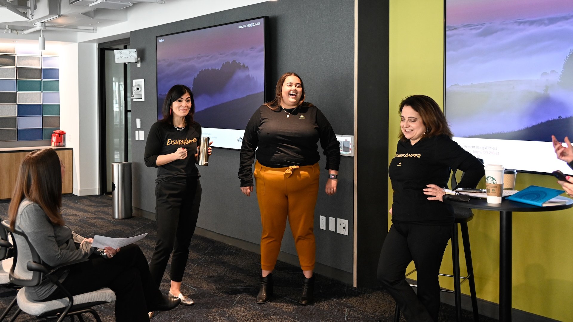 Group of people in office setting, some standing and some sitting, one is laughing.