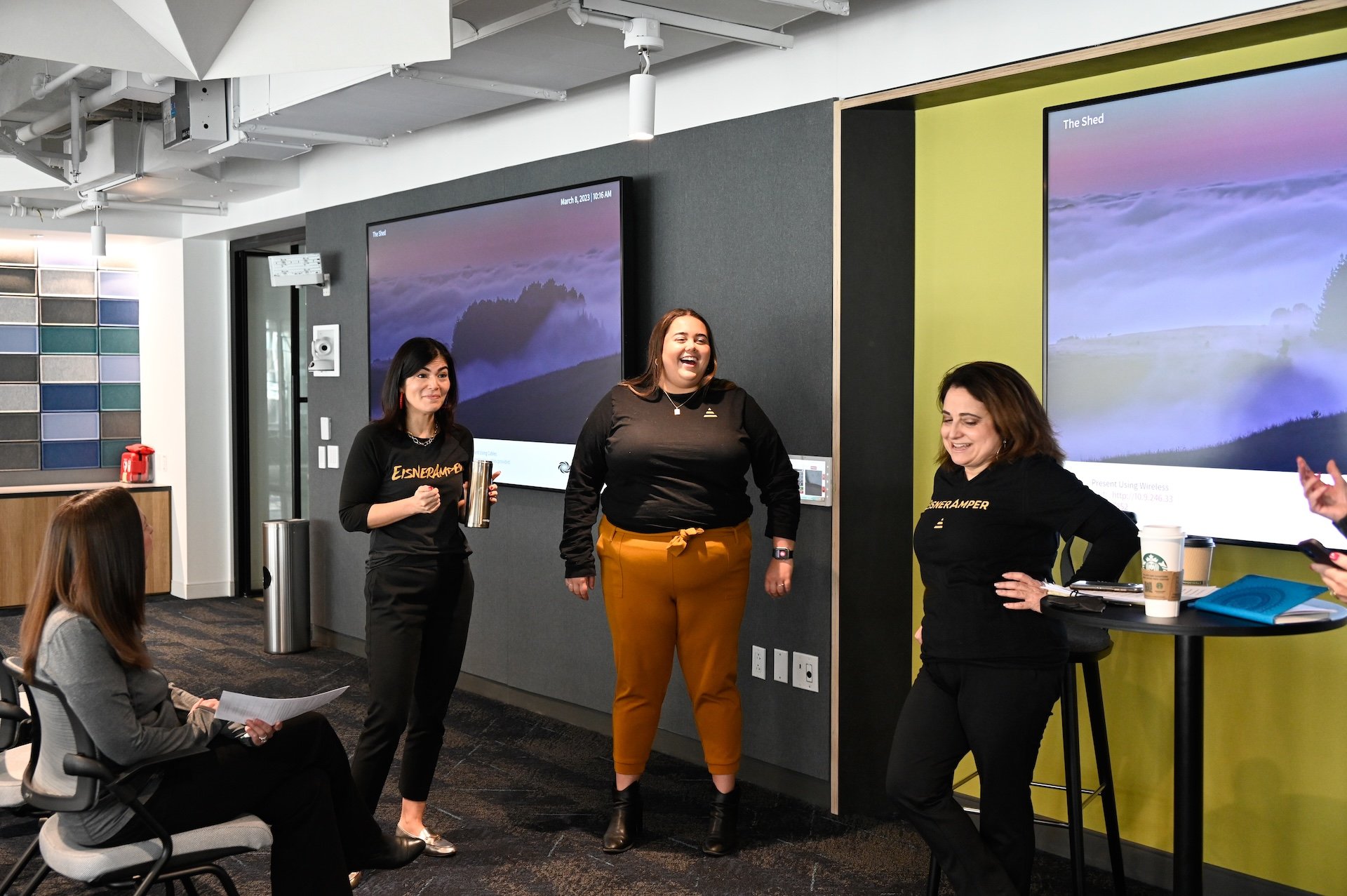 Group of people in office setting, some standing and some sitting, one is laughing.